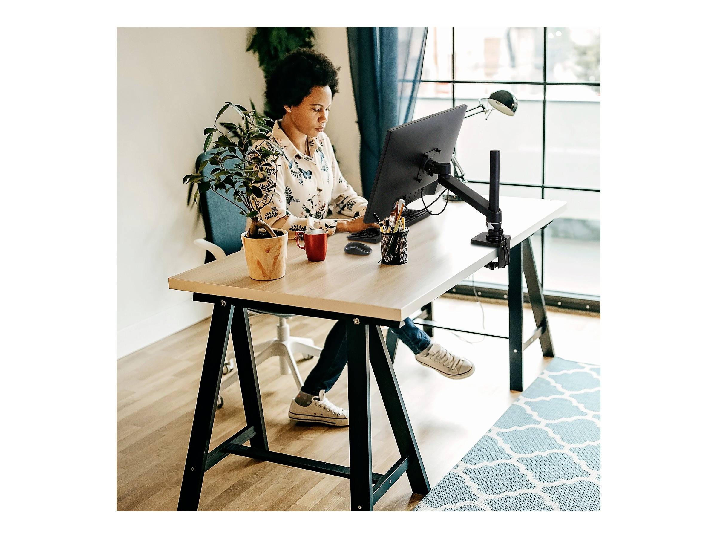 Osoba pracuje w skupieniu przy monitorze komputerowym w jasnym biurze z dużym oknem w tle.