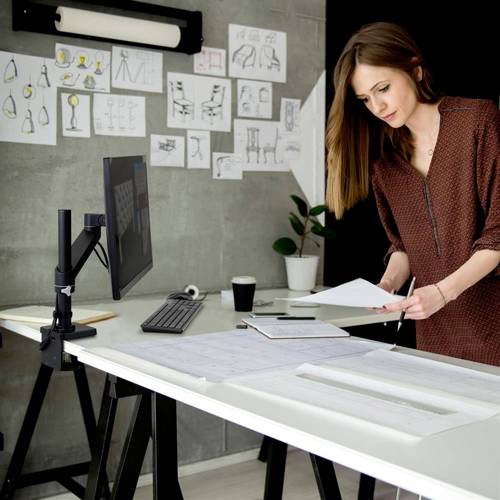 Kobieta stoi przy biurku z monitorem i sprawdza plany. Na ścianie wiszą rysunki i projekty lamp oraz mebli.