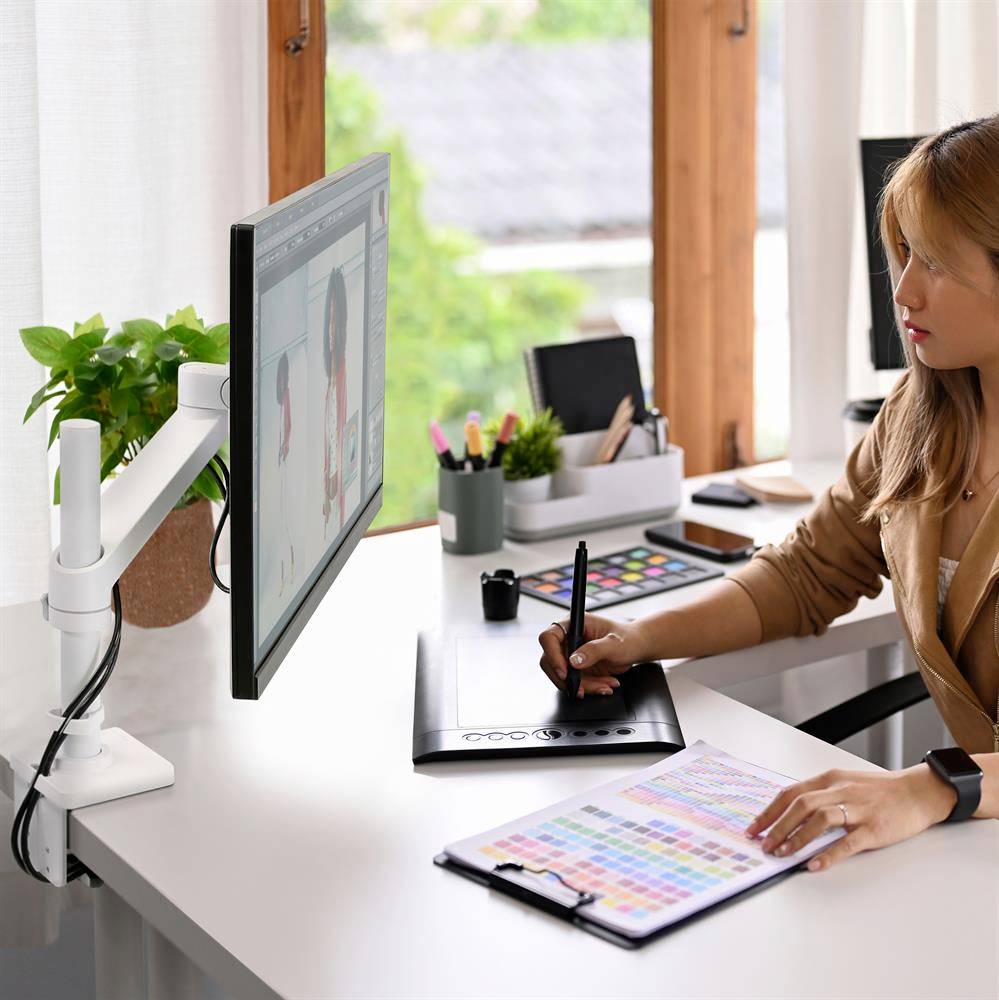 Kobieta pracuje kreatywnie na komputerze przy użyciu tabletu graficznego, otoczona materiałami biurowymi. W tle okno wpuszcza światło dzienne.