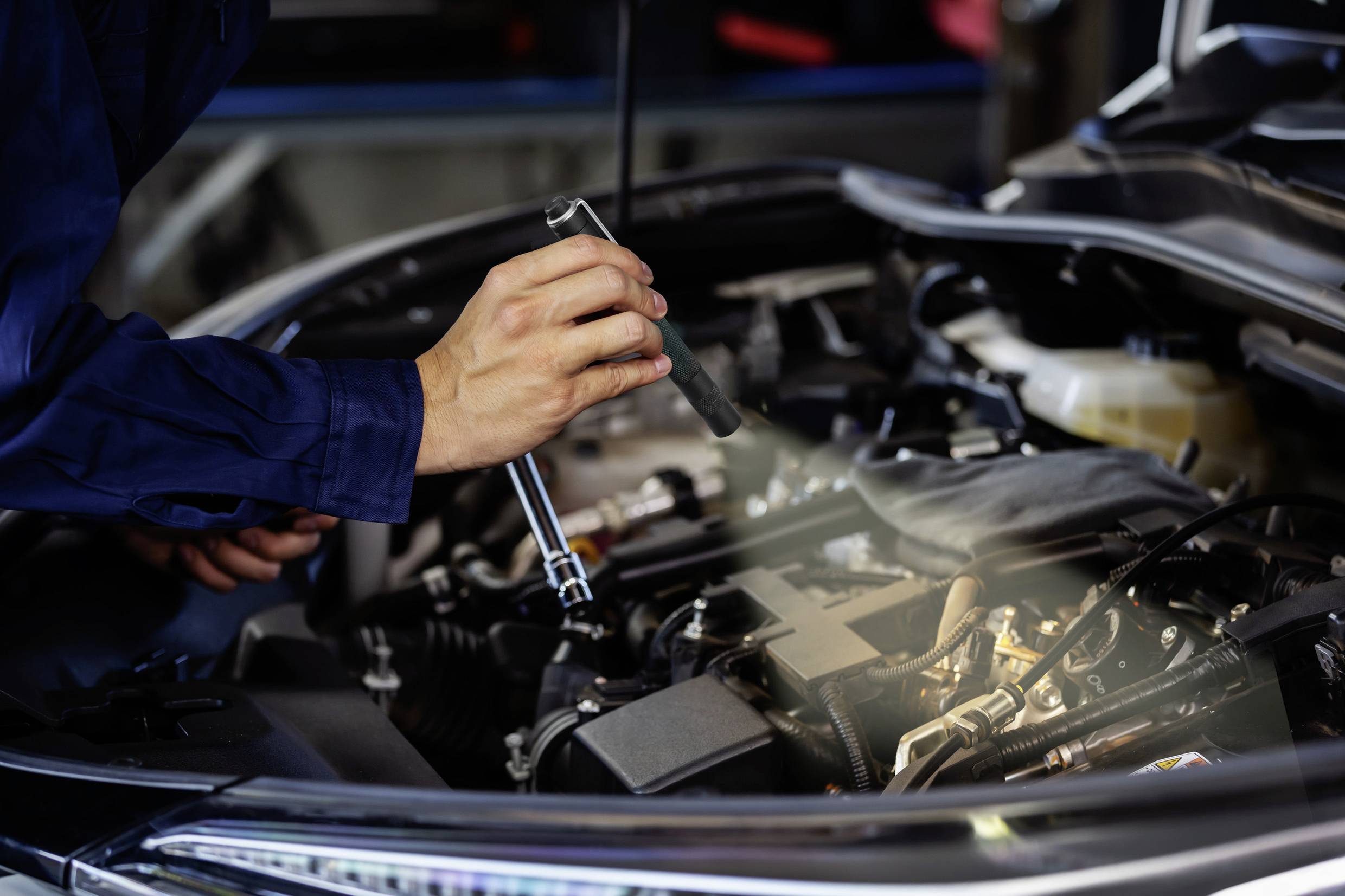 Mechanik sprawdza silnik samochodu latarką, trzymając w drugiej ręce klucz w warsztacie.