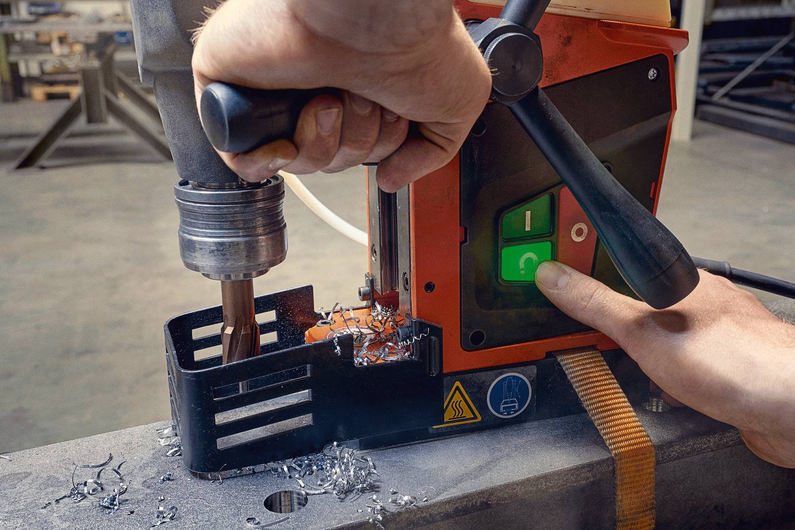 Osoba obsługuje przemysłową wiertarkę, aby wywiercić otwór w kawałku metalu. Ręce trzymają wiertło i maszynę. Widoczne są przyciski sterujące.