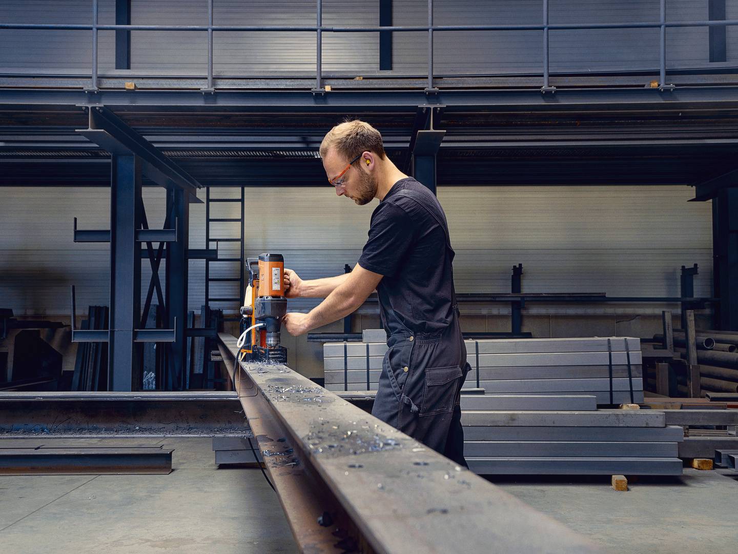 Mężczyzna w odzieży roboczej używa wiertarki do wykonywania otworów w długim elemencie metalowym w przemysłowym warsztacie.
