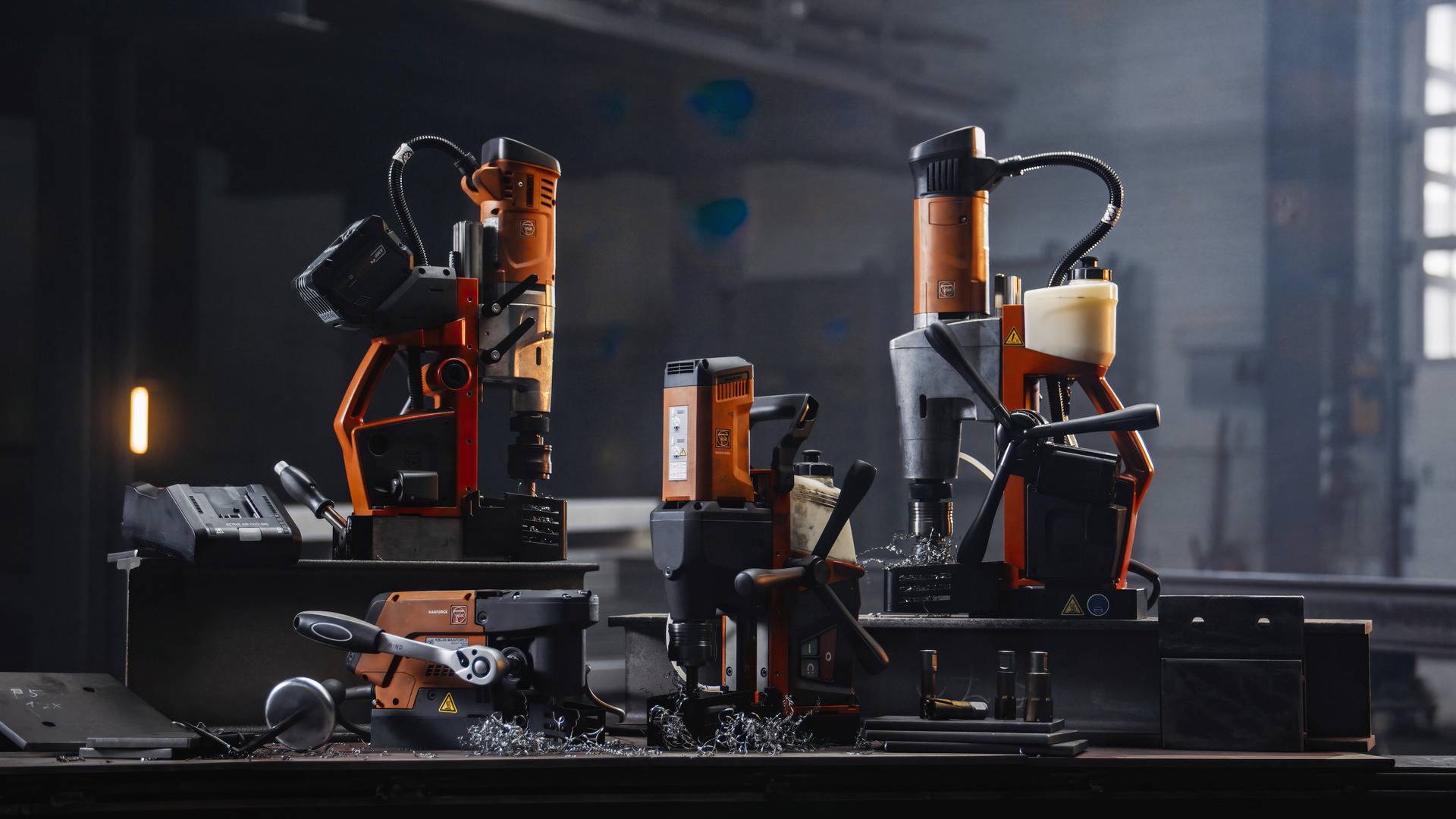 Różne wiertarki stoją na stole warsztatowym w warsztacie. Wokół urządzeń rozrzucone są metalowe wióry. W tle widać okno warsztatu.