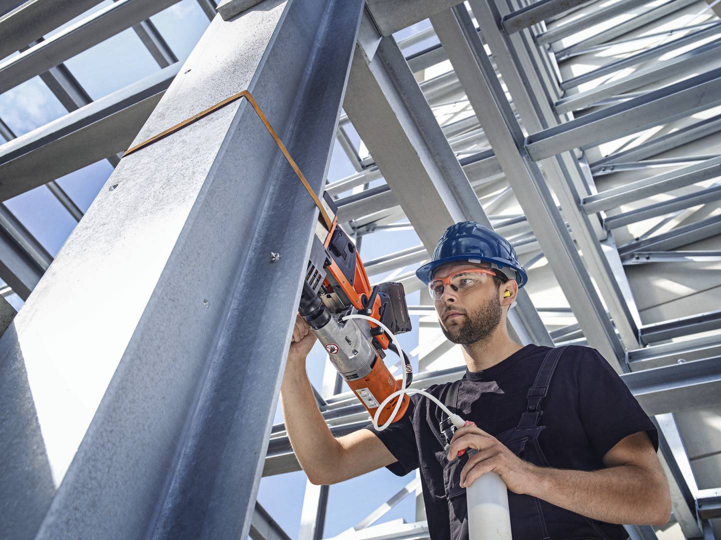 Robotnik budowlany w kasku ochronnym i okularach pracuje z wiertnicą przy stalowej konstrukcji na terenie budowy.