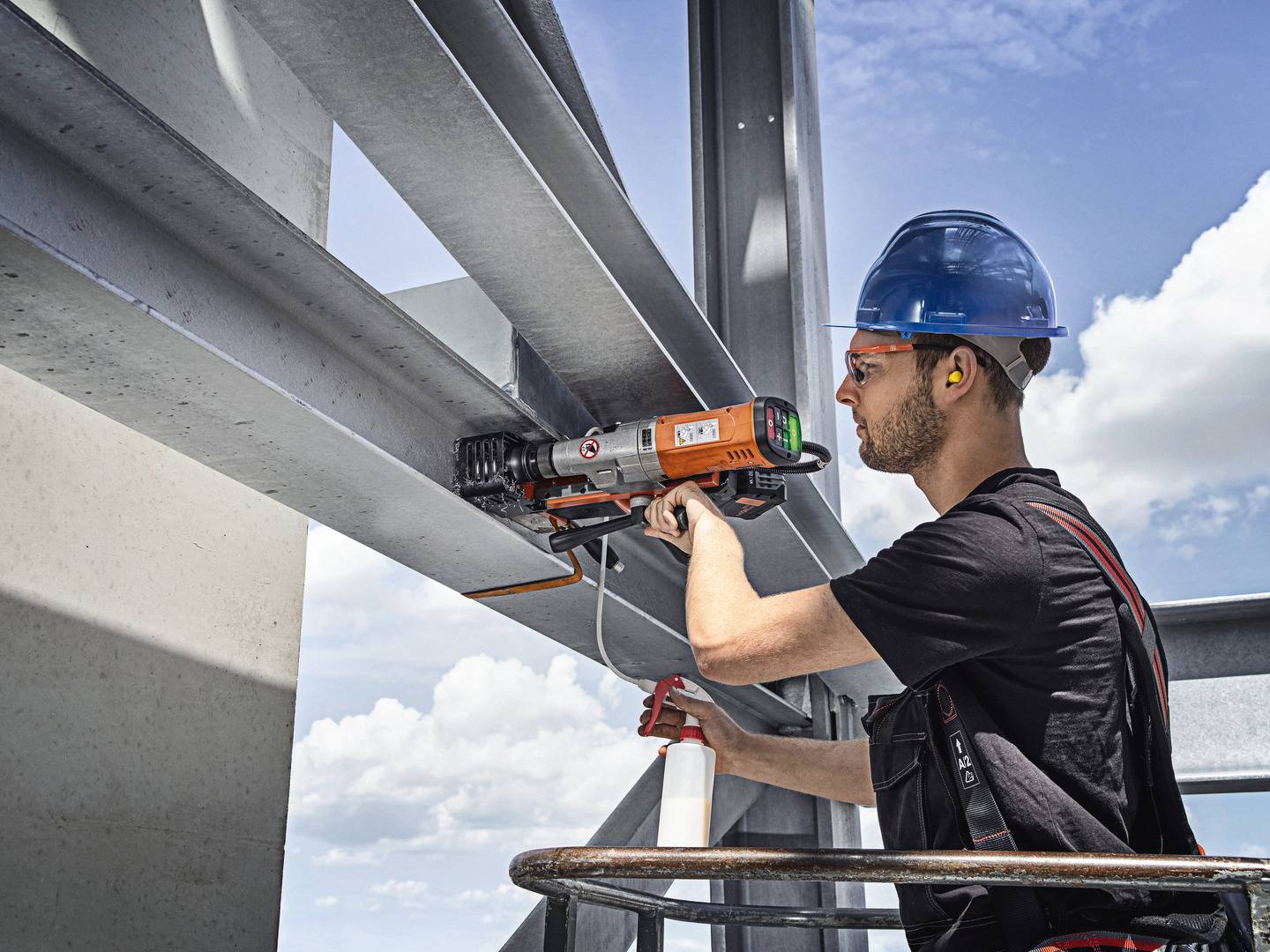Robotnik budowlany w kamizelce ochronnej skręca stalowe belki przy pomocy elektronarzędzia. W tle widać błękitne niebo.