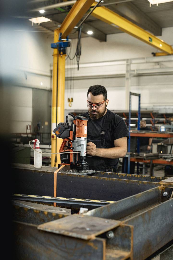 Mężczyzna pracuje z maszyną w warsztacie. Nosi okulary ochronne i jest całkowicie skupiony na swojej pracy. W tle widać półki i narzędzia.