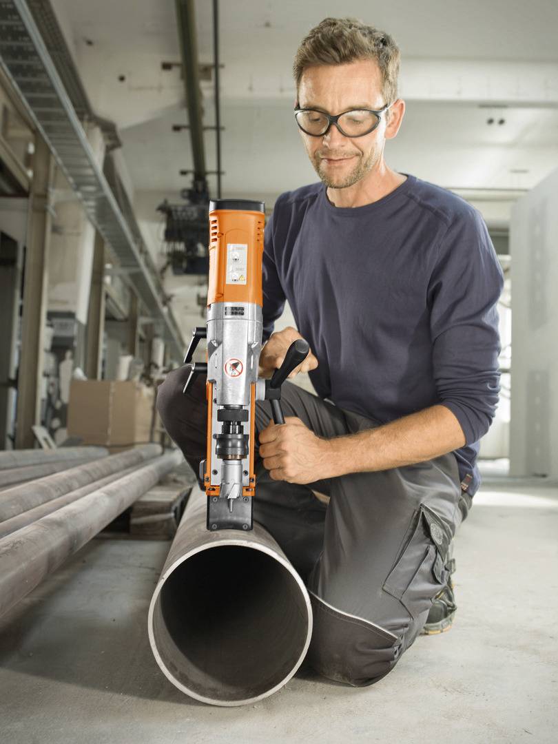 Mężczyzna w okularach ochronnych wierca dużą wiertarką w rurę metalową w warsztacie.