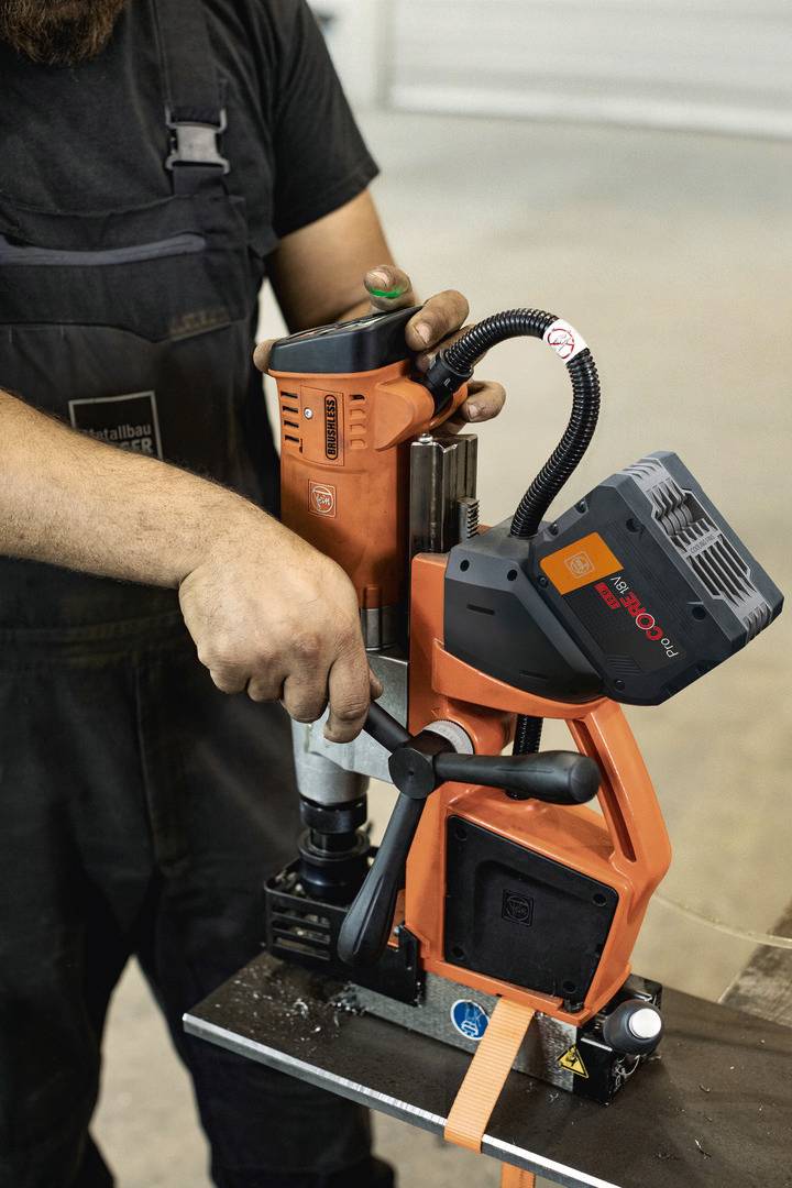 Osoba używa magnetycznej wiertarki na płycie metalowej. Fokus jest na maszynie i procesie wiercenia.