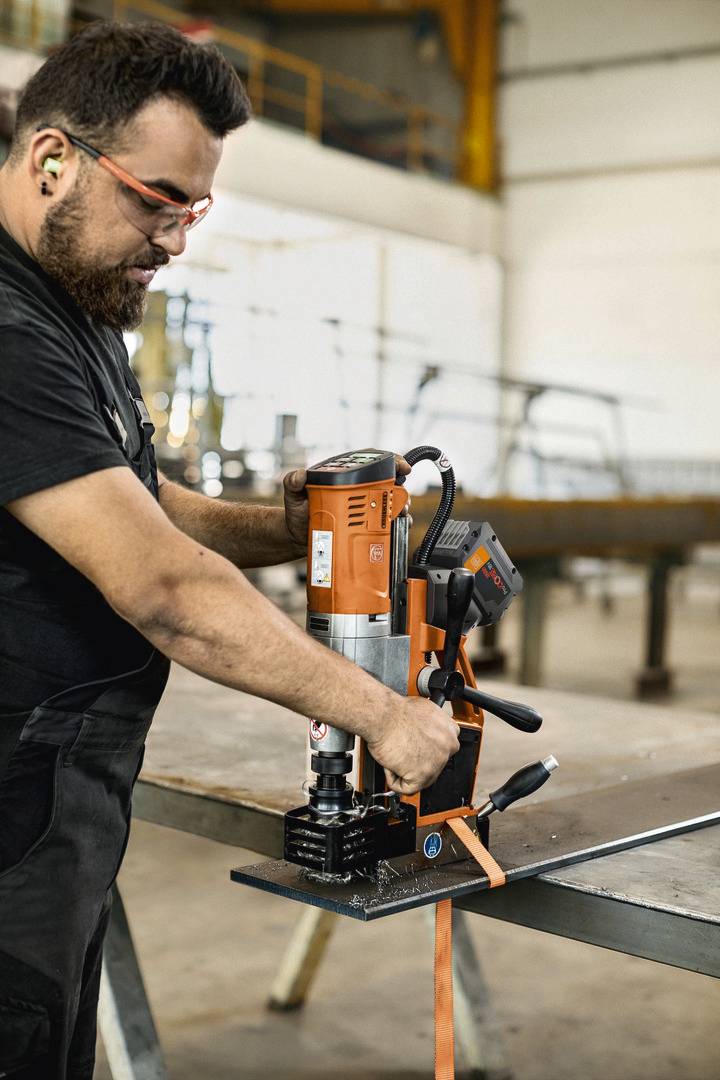 Mężczyzna w okularach ochronnych i zatyczkach do uszu obsługuje dużą wiertarkę na metalowym stole w warunkach warsztatowych.