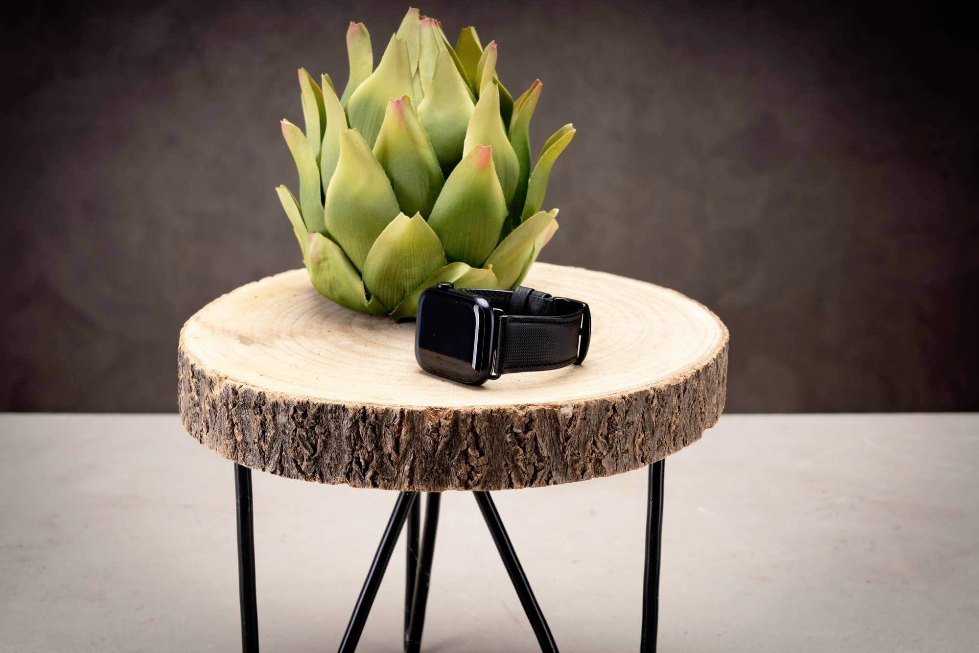 Smartwatch z czarnym paskiem leży na stylowym, okrągłym drewnianym stole przed zielonym elementem dekoracyjnym przypominającym karczocha.