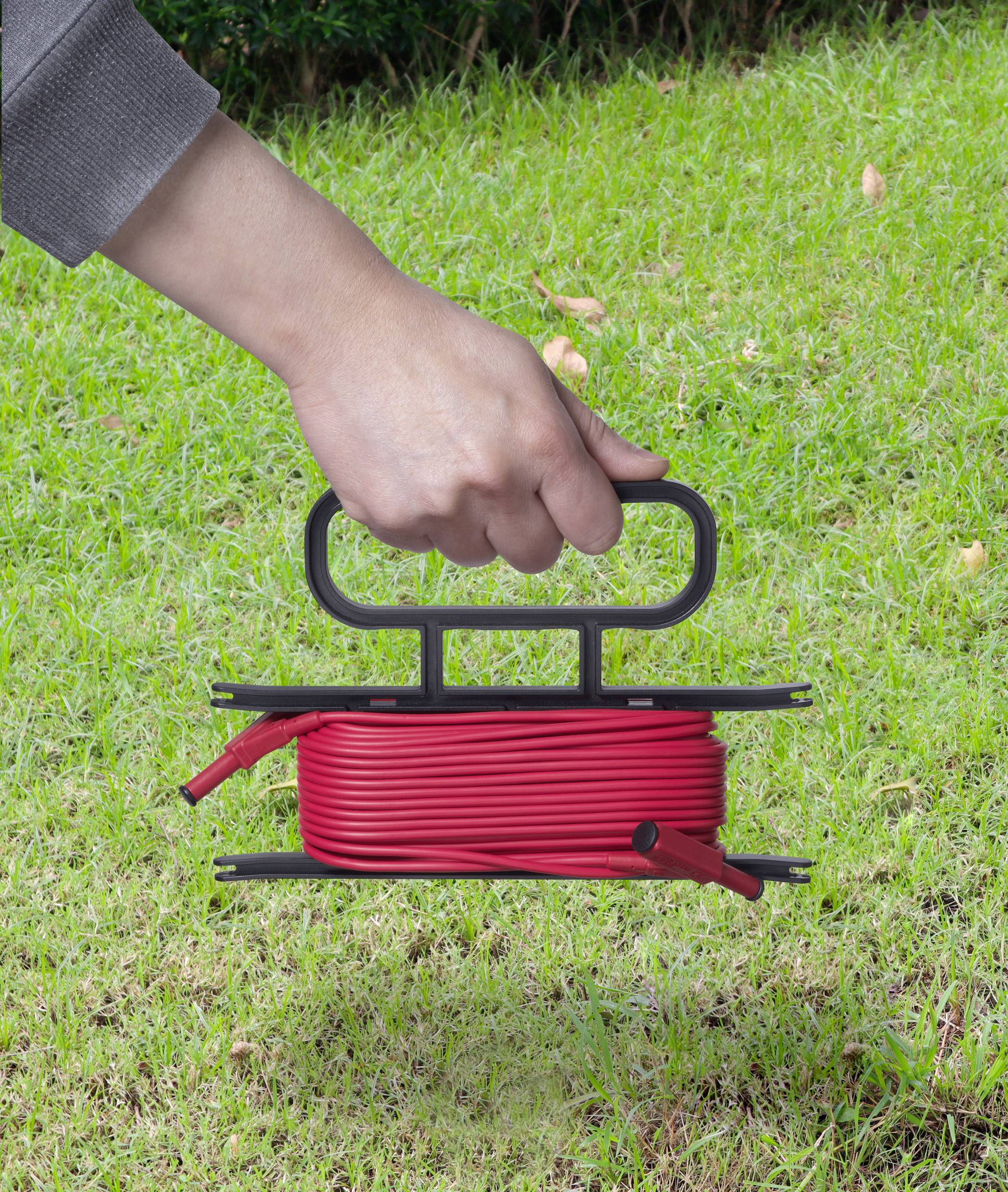 Osoba trzyma czerwoną, zwin��tą rolkę kabla z uchwytem na zielonej trawie.