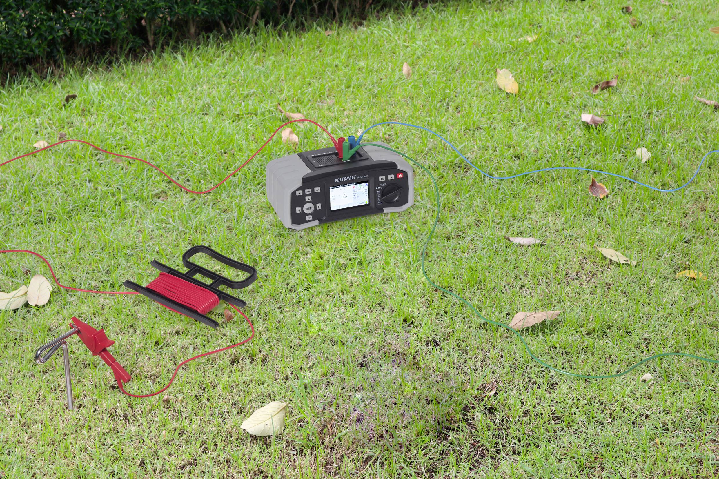 Cyfrowy miernik gruntowy leży na zielonej trawie, podłączony wieloma kablami i uziemiaczem. Liście są rozrzucone na ziemi.