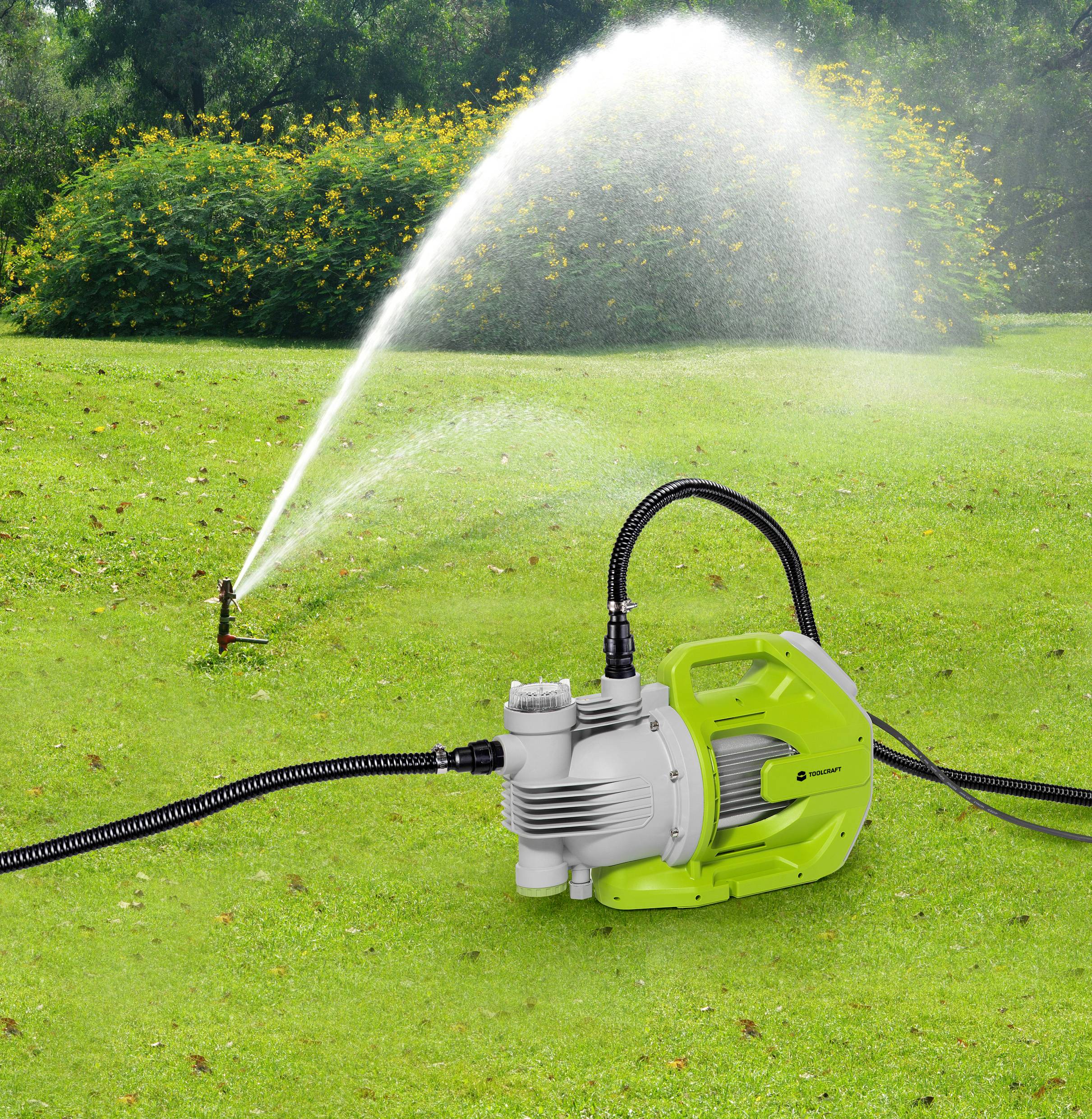 Zielona pompa wodna na trawniku, podłączona do węża, który kieruje wodę do zraszacza, równomiernie nawadniającego ogród.