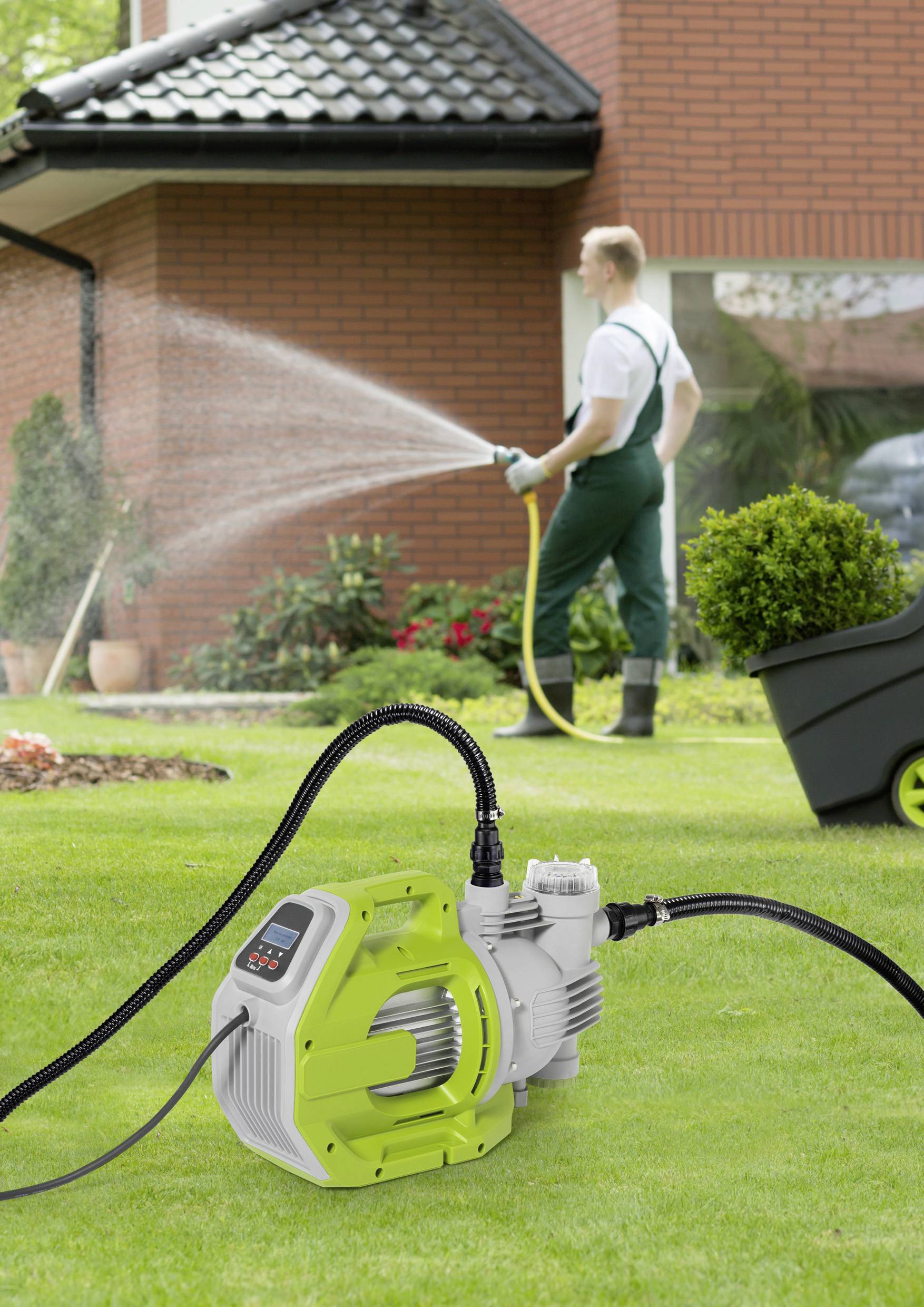 Ogrodnik nawadnia trawnik za pomocą węża, podczas gdy na pierwszym planie widoczna jest elektryczna pompa wodna.