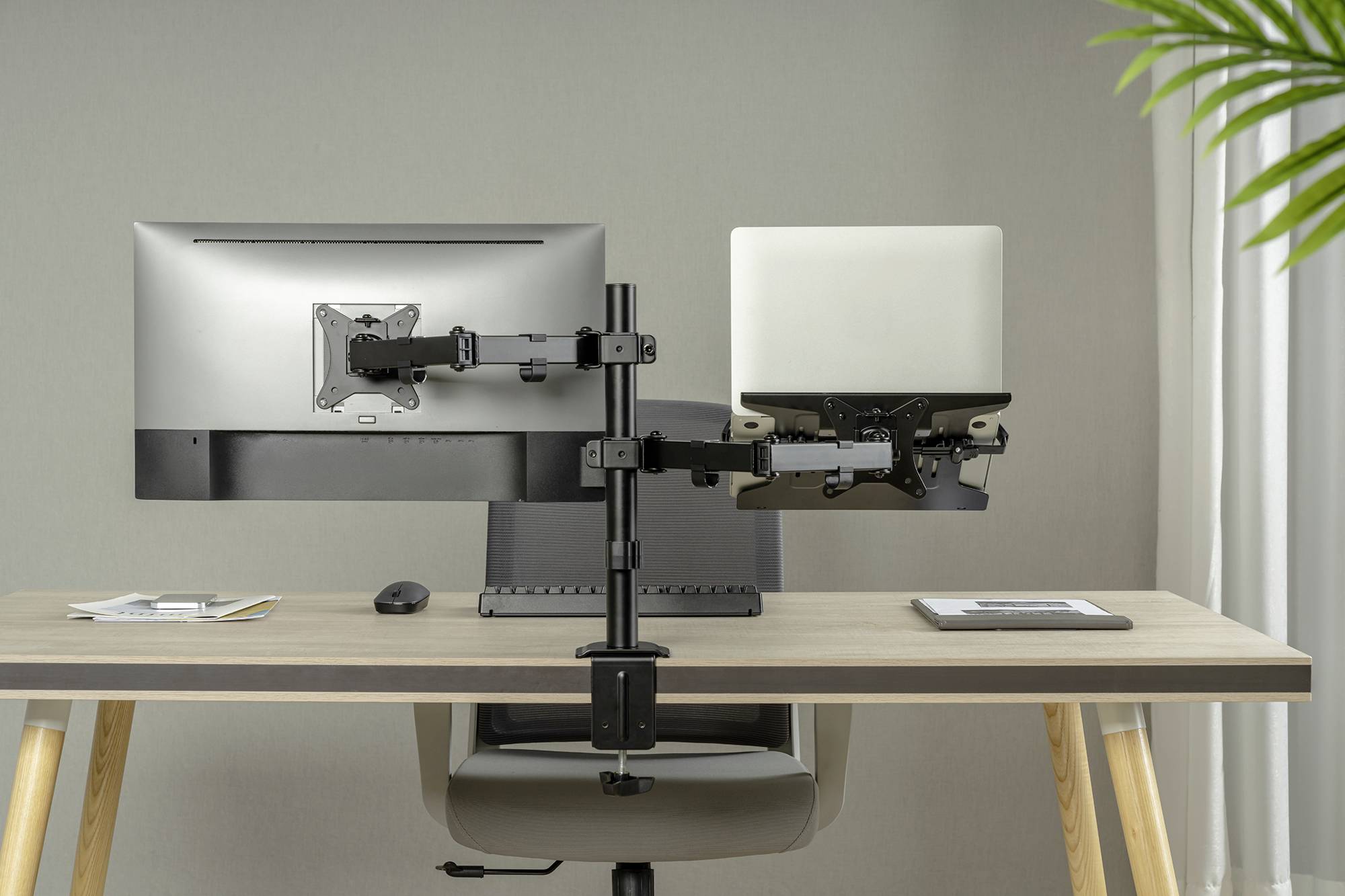 Dwa monitory zamocowane na podwójnym ramieniu na biurku, jeden wyświetla ekran laptopa, obok znajdują się artykuły biurowe. W tle neutralna ściana.