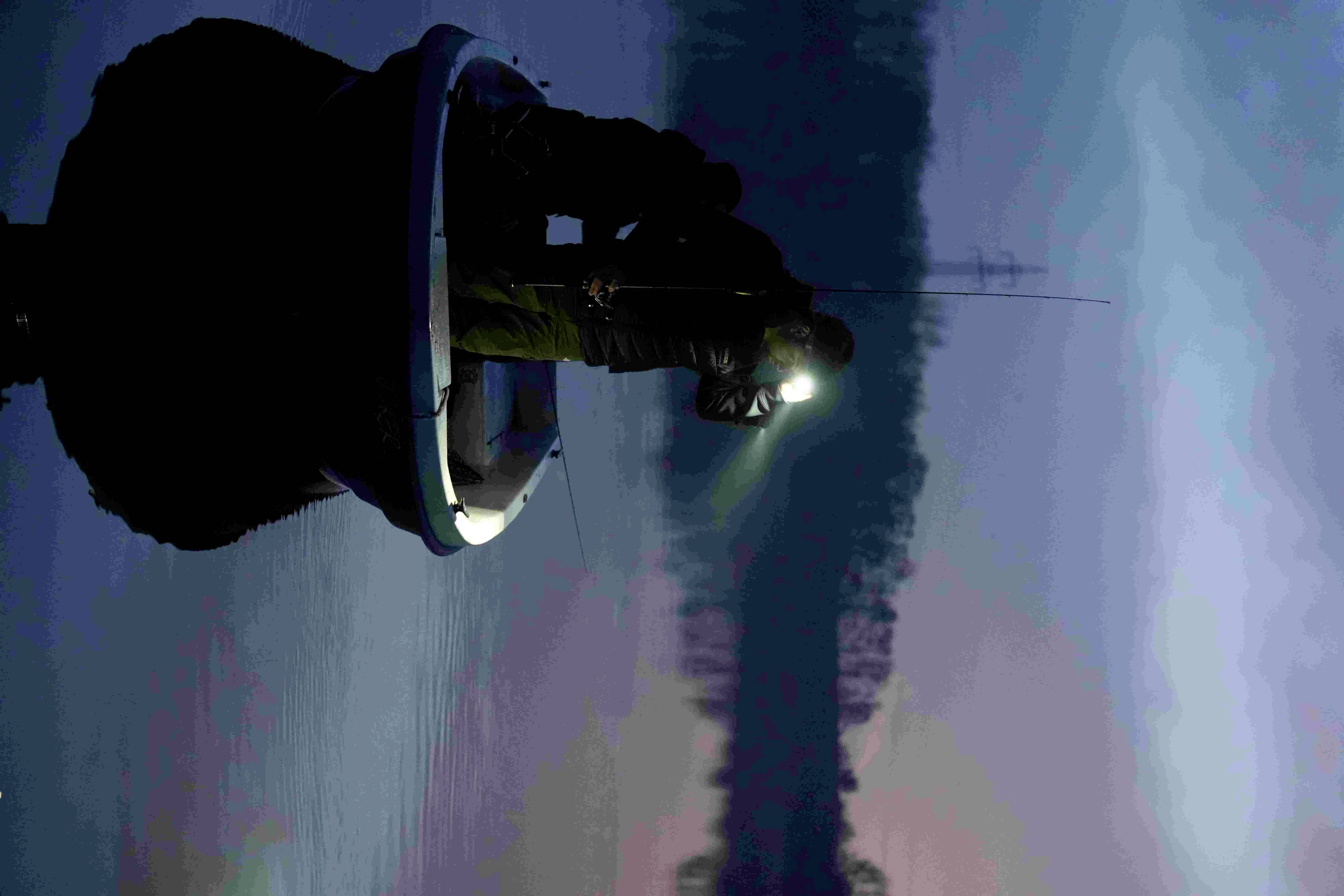 Dwie osoby łowią ryby nocą na małej łodzi na spokojnym jeziorze. Jedna z nich trzyma latarkę, która oświetla wodę.