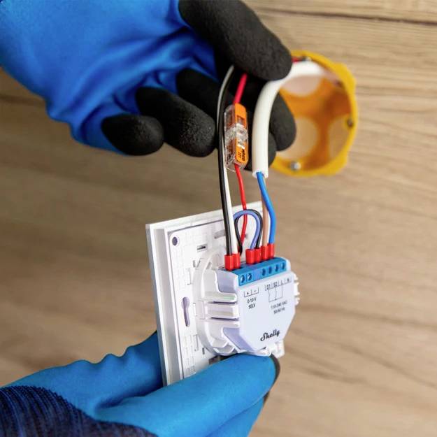 Osoba montuje urządzenie elektroniczne na ścianie. Nosi niebieskie rękawice i łączy kolorowe przewody z gniazdkiem elektrycznym.