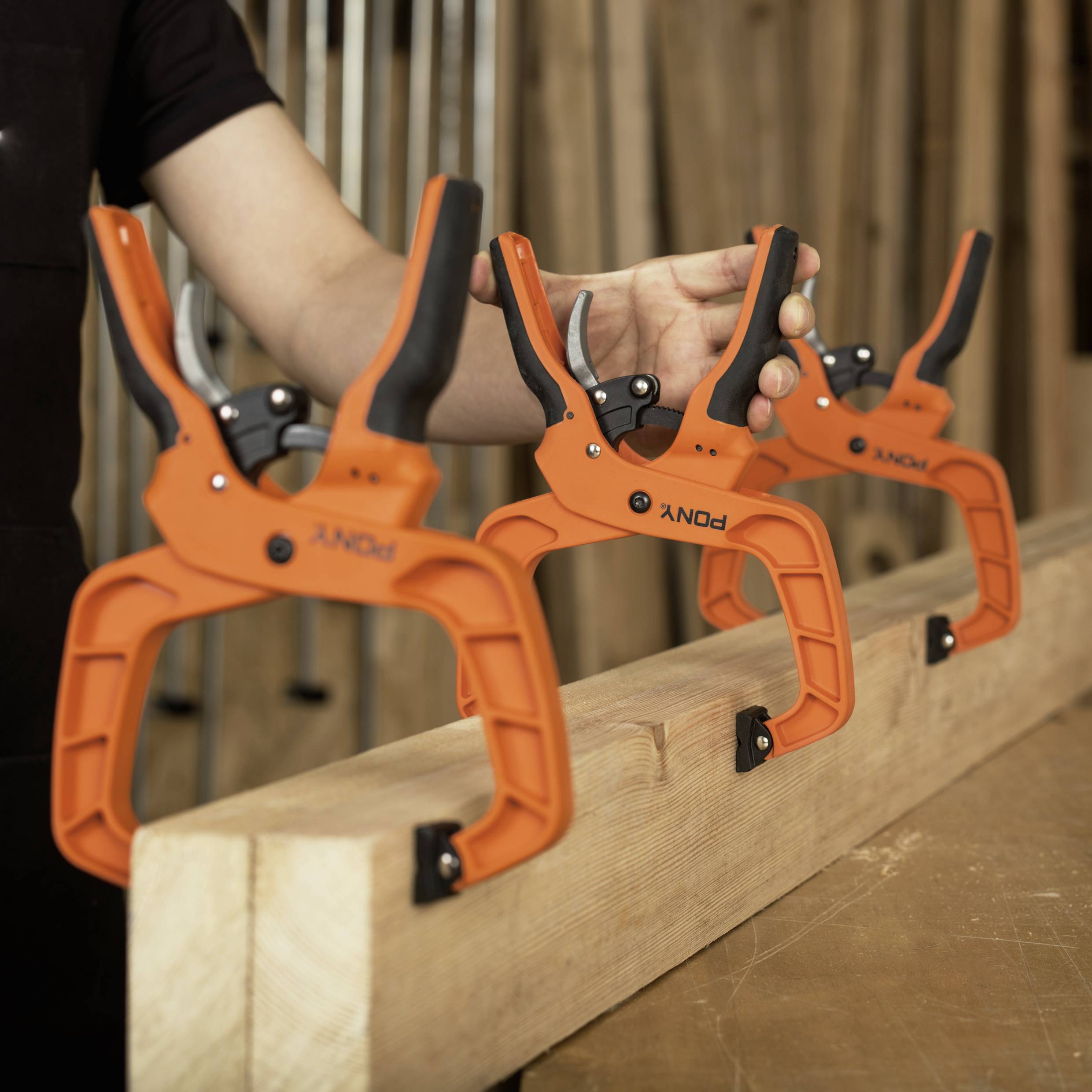 Trzy pomarańczowe zaciski przytrzymują kawałek drewna. Człowiek reguluje zaciski. Są używane do prac stolarskich.