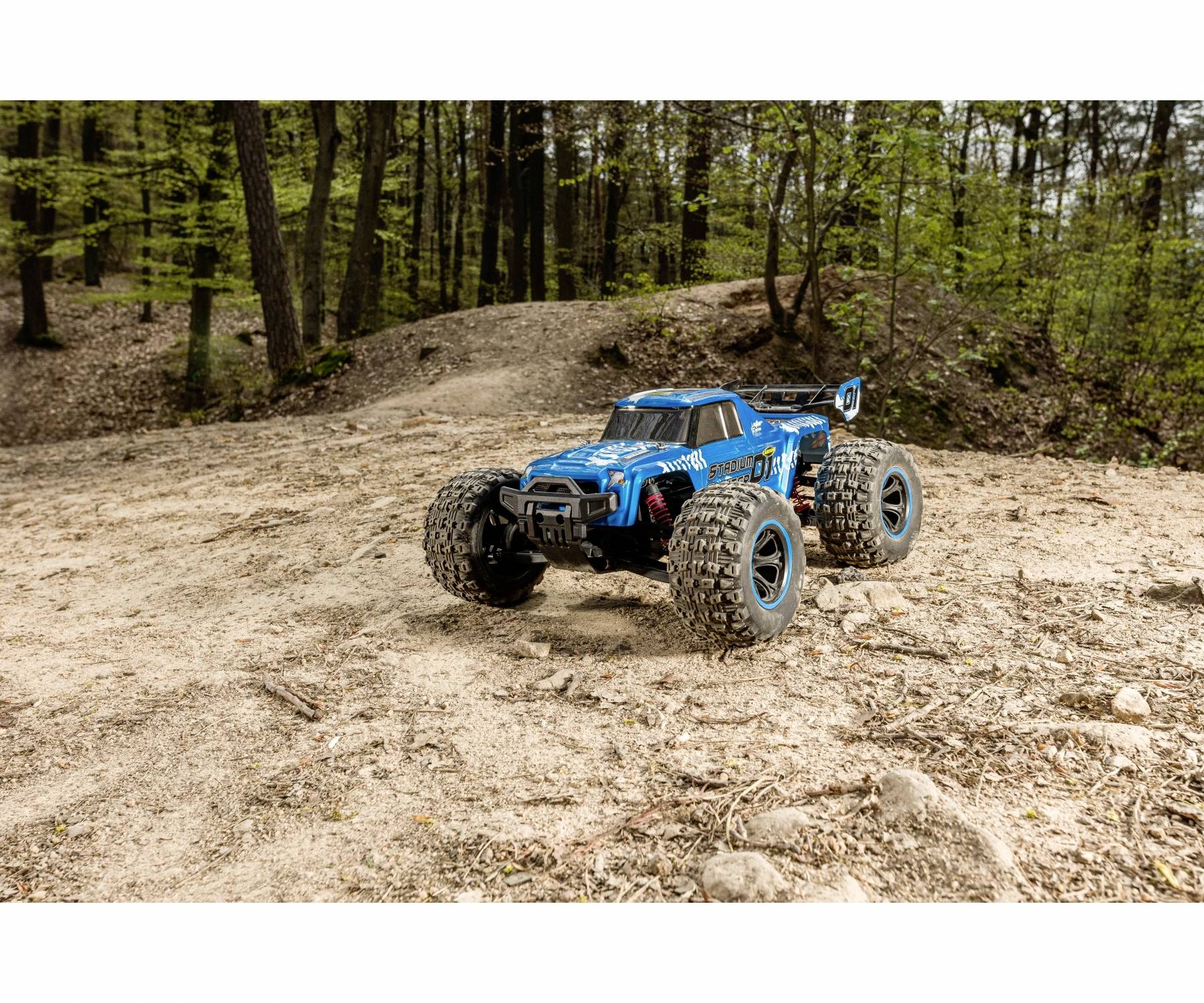 Niebieskie, zdalnie sterowane auto jedzie piaszczystą drogą w lesie. Drzewa otaczają scenę, podczas gdy auto znajduje się na pierwszym planie.