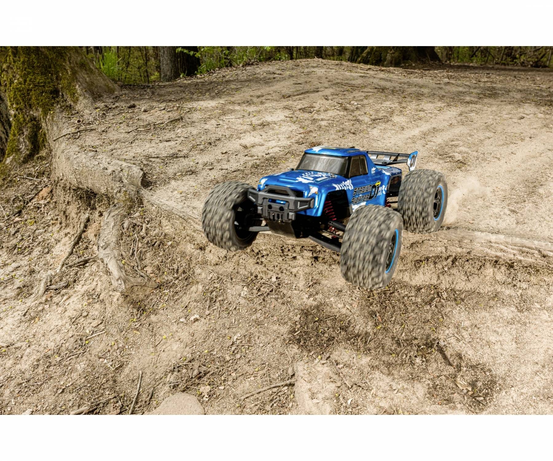 Niebieski zdalnie sterowany samochód terenowy skacze przez piaszczysty pagórek w leśnym terenie.