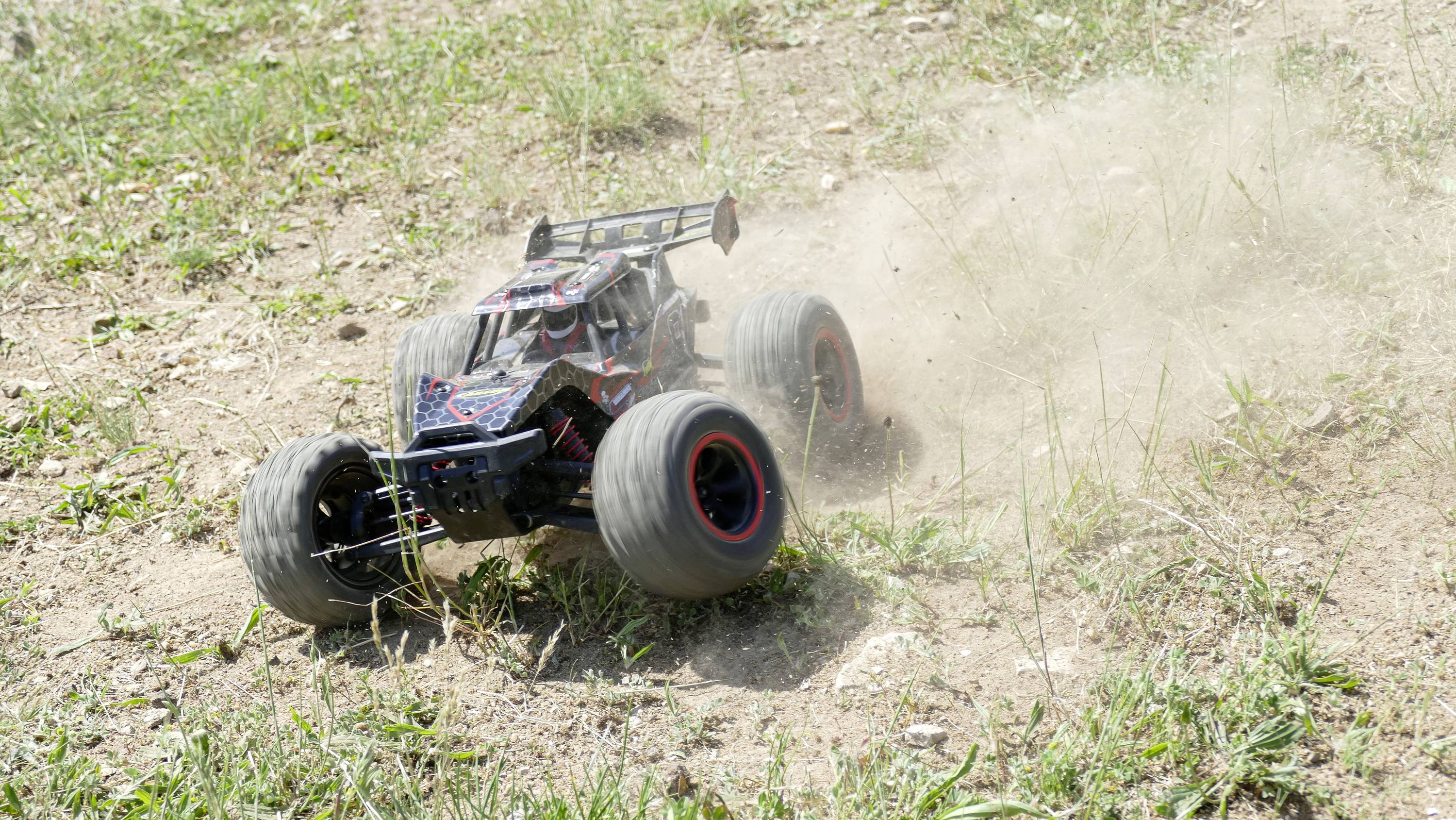 Zdalnie sterowany samochód pędzi z dużą prędkością po pylistej, pokrytej trawą przestrzeni. Za samochodem unosi się kłęby kurzu.