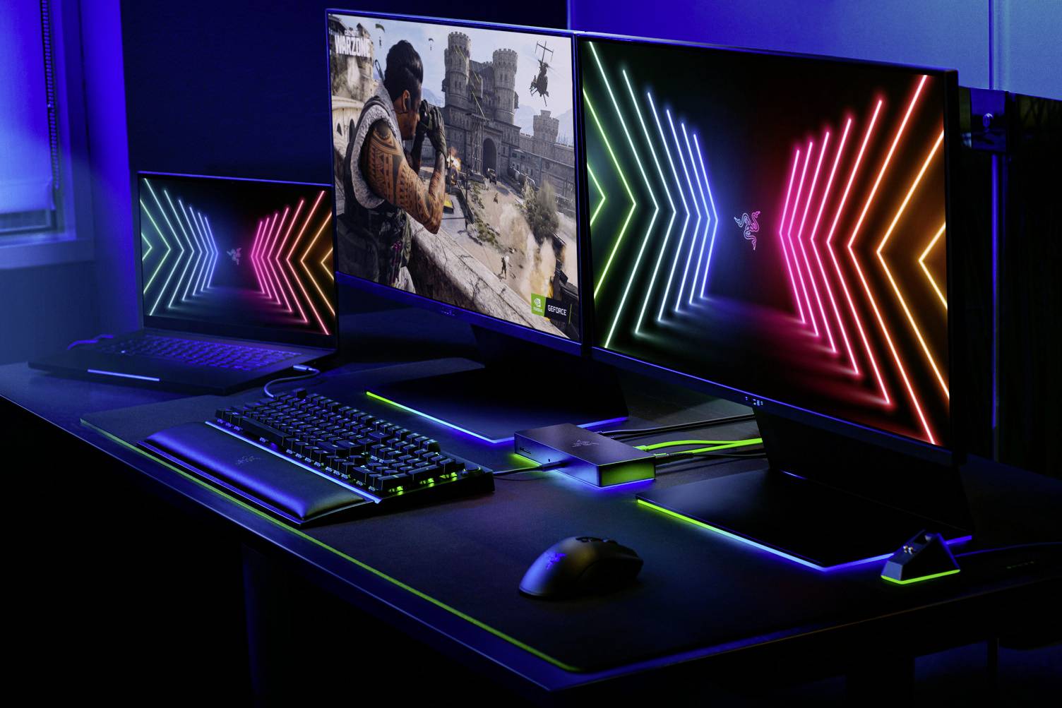 Dwa monitory gamingowe na biurku; lewy wyświetla grę, prawy kolorowe wzory świetlne. Klawiatura i mysz pośrodku.