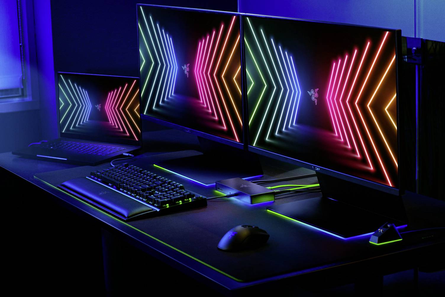 Zestaw do gier z dwoma monitorami oraz klawiaturą i myszą w świecących kolorach neonowych, tworzący futurystyczną atmosferę.