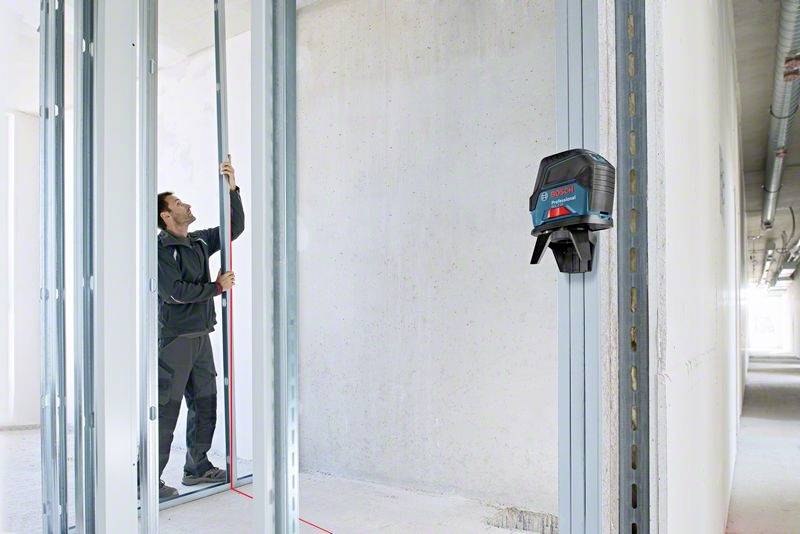 Rzemieślnik mierzy odległość między ściankami z płyt kartonowo-gipsowych za pomocą laserowego urządzenia pomiarowego. W tle widać plac budowy.