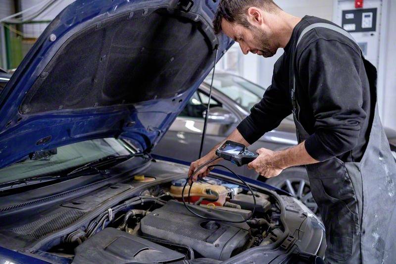 Mechanik diagnozuje problem w komorze silnika samochodu za pomocą ręcznego urządzenia. Stoi skupiony obok pojazdu.