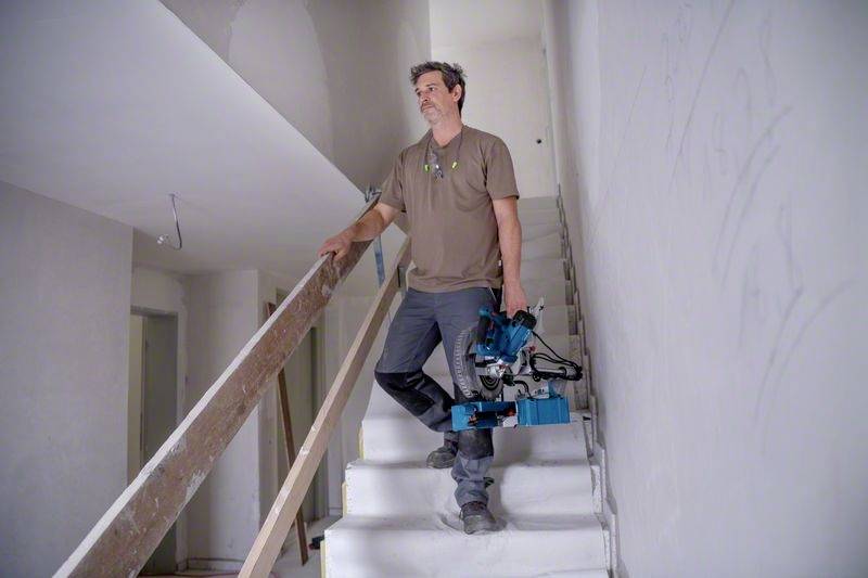 Mężczyzna stoi na schodach w niedokończonym budynku, trzymając elektryczną piłę. Pomieszczenie ma otynkowane ściany i tymczasową balustradę.