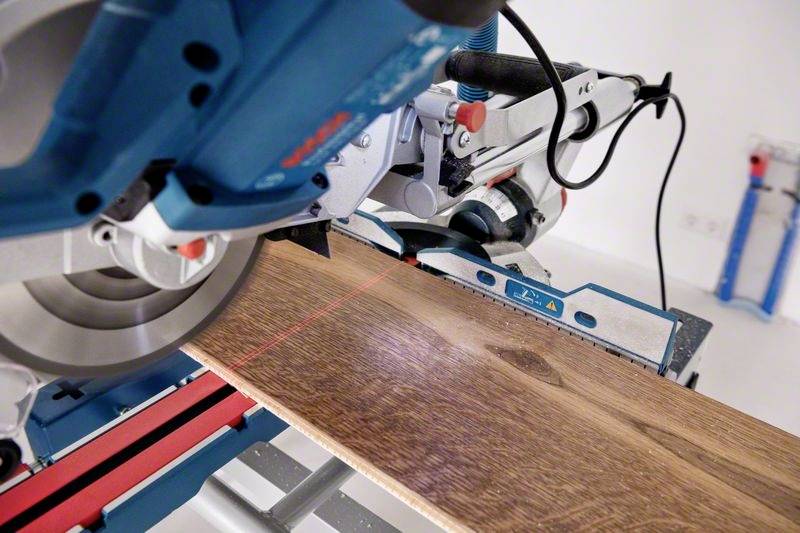 Ręczna piła tarczowa tnie deskę drewnianą. Czerwona wiązka laserowa wskazuje linię cięcia. W tle widoczne są narzędzia.