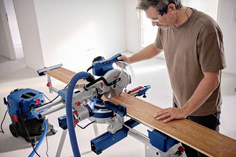 Mężczyzna przecina długą deskę drewnianą piłą ukośnicową w warsztacie. Do piły podłączony jest odkurzacz pyłu.
