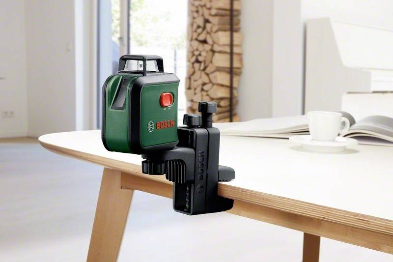 Zielony laser Boscha zamocowany na stole, celuje czerwonym promieniem laserowym. Obok lasera stoi biały kubek do kawy.