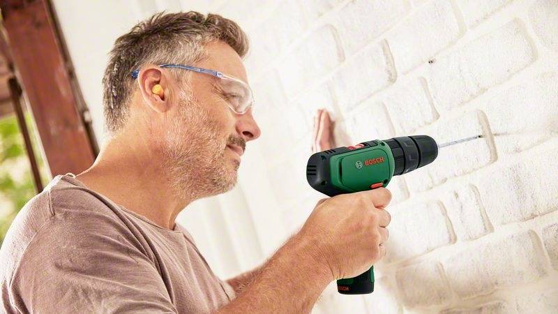 Mężczyzna wierci dziurkę w białej ścianie z cegły za pomocą zielonego, bezprzewodowego wkrętarki akumulatorowej. Na oczach ma okulary ochronne, a w uszach - zatyczki.