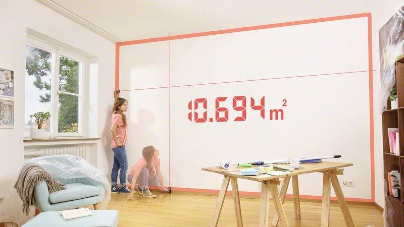Dziecko używa laserowego miernika przy białej ścianie w jasnym pokoju. Na wyświetlaczu widnieją wymiary ściany wynoszące '10,694 m²'.