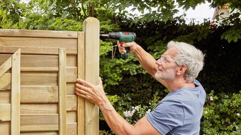Starszy mężczyzna mocuje drewniany płot w ogrodzie za pomocą akumulatorowego wkrętarki. Ma na sobie niebieską koszulkę i pracuje z dużym skupieniem.