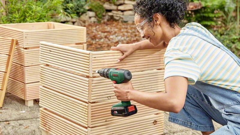 Osoba w okularach ochronnych używa wiertarki do montażu drewnianej skrzyni na wolnym powietrzu. W tle widoczne są rośliny i kamienie.