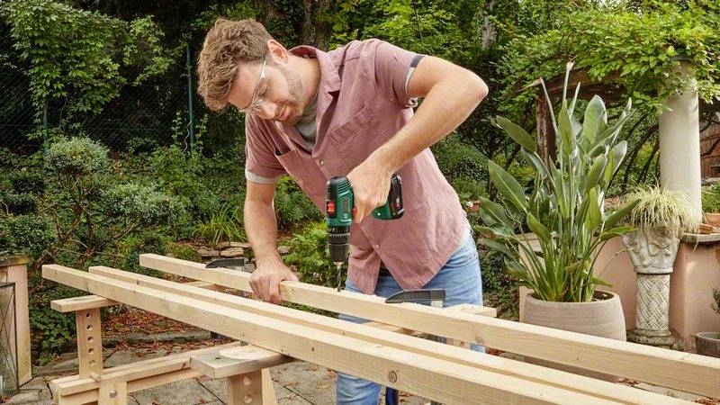 Mężczyzna wierca dziurę w desce drewnianej na wolnym powietrzu. Nosi okulary ochronne i używa zielonej wiertarki. W tle widoczne są rośliny.