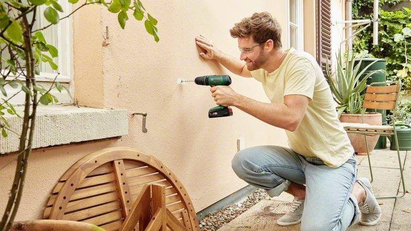 Mężczyzna klęczy na tarasie i wierci wiertarką akumulatorową otwór w zewnętrznej ścianie domu, aby coś zamocować.