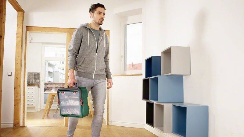 Mężczyzna w stroju casual niesie skrzynkę narzędziową przez pomieszczenie z kolorowymi regałami ściennymi.