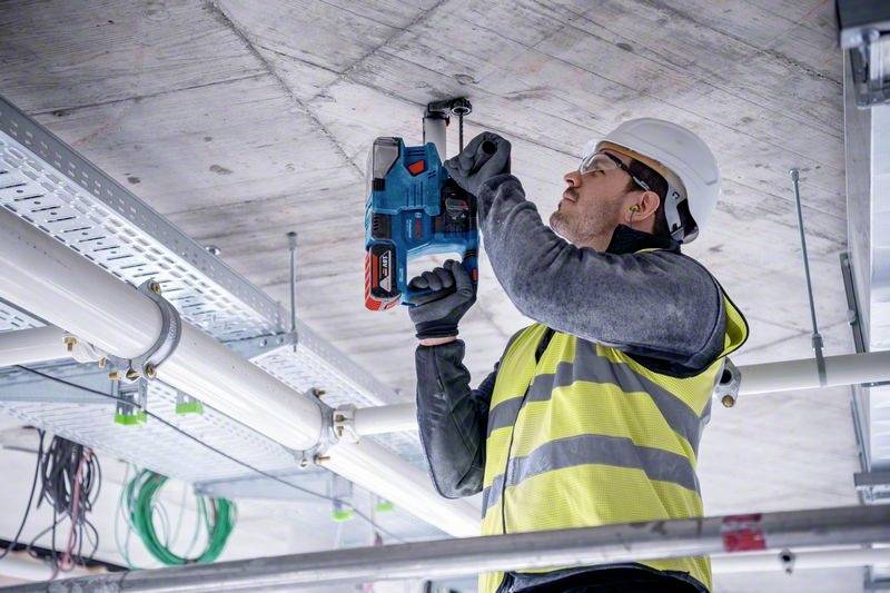 Robotnik na budowie w kasku i kamizelce odblaskowej, wiercący elektrycznym narzędziem w suficie. W tle widoczne są przewody i kable.