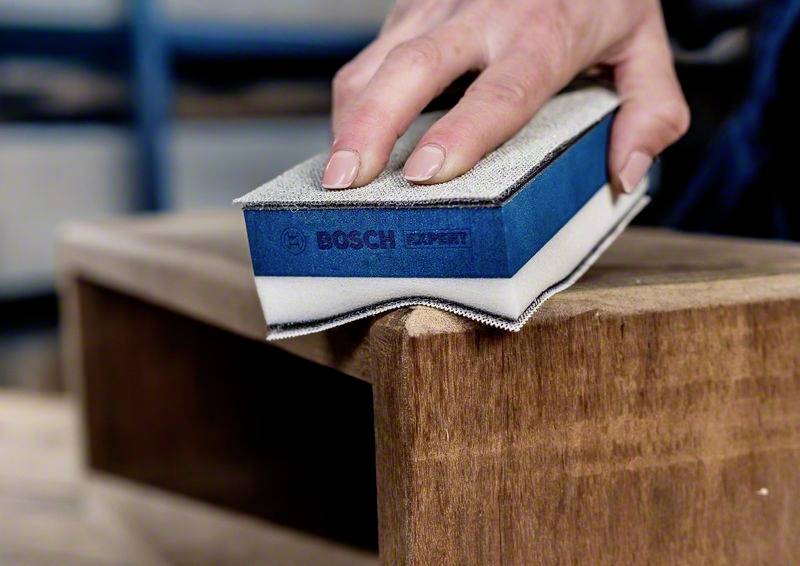 Ręka szlifuje krawędź drewna blokiem szlifierskim Boscha. Powierzchnia deski drewnianej jest wygładzana.
