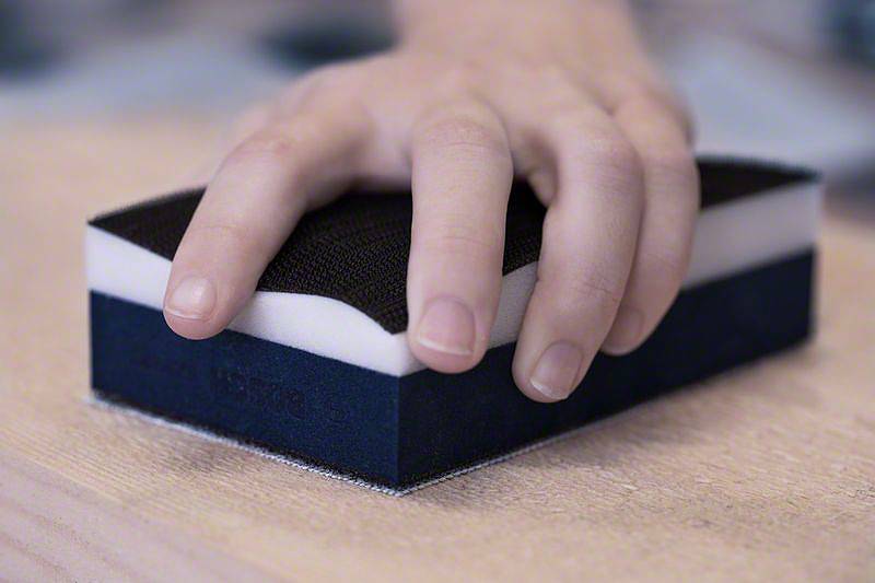 Jedna ręka trzyma gąbkę ścierną używaną na powierzchni drewna. Gąbka posiada czarną i białą warstwę.