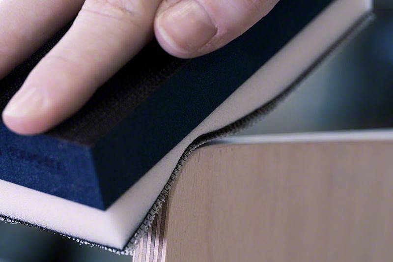 Dłoń szlifuje krawędź drewna gąbką ścierną, aby wygładzić powierzchnię.