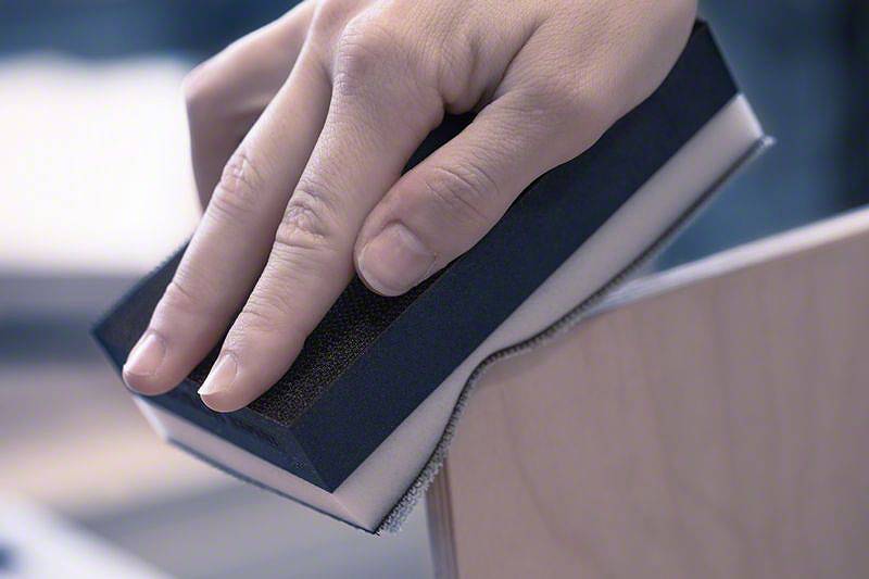 Dłoń szlifuje krawędź elementu drewnianego blokiem szlifierskim.