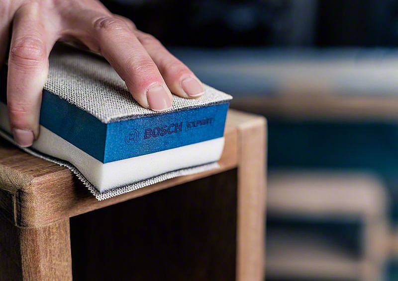 Jedna ręka trzyma gąbkę ścierną i szlifuje krawędź drewnianego elementu meblarskiego. Gąbka nosi logo znanej marki.