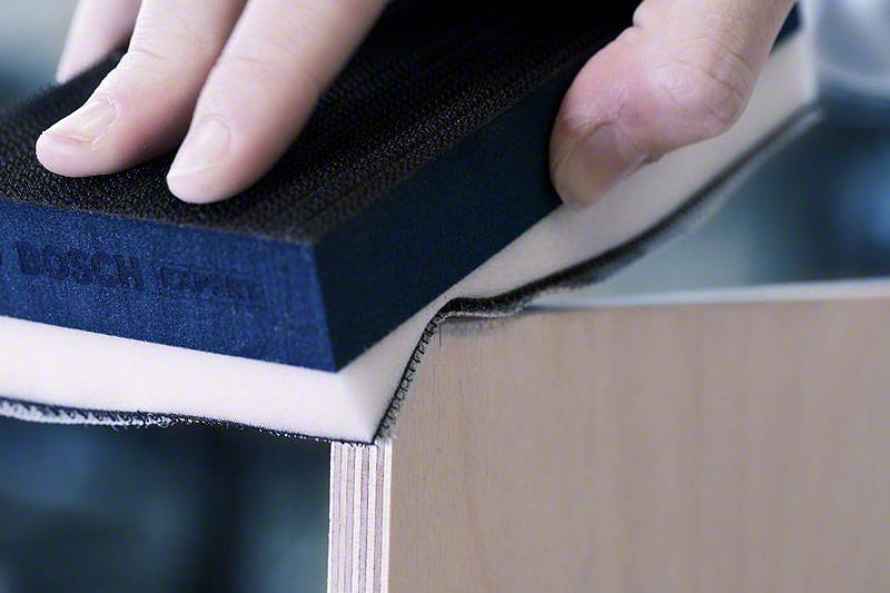 Ktoś wyrównuje krawędź deski drewnianej za pomocą bloku szlifierskiego. Fokus skierowany jest na dłoń i blok szlifierski.