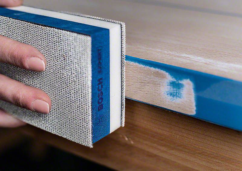 Osoba szlifuje krawędź deski drewnianej za pomocą padu szlifierskiego, aby wygładzić powierzchnię. Krawędź jest oznaczona na niebiesko.