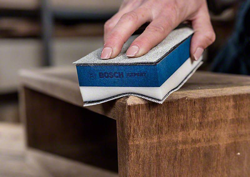 Ręka szlifuje drewno gąbką ścierną Bosch. Nacisk położony jest na precyzję szlifowania oraz charakterystyczny niebieski kolor gąbki.