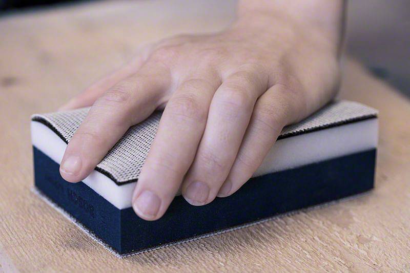 Ręka trzyma blok szlifierski, który przesuwa się po powierzchni drewna, obrabiając ją.